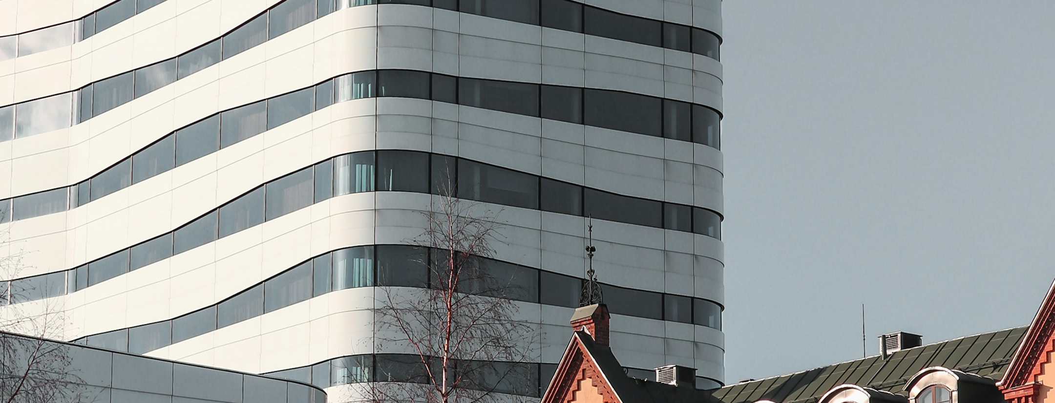 Modern byggnad med böljande fasad i vitt och glas, framför äldre hus med tegel och grönt tak.