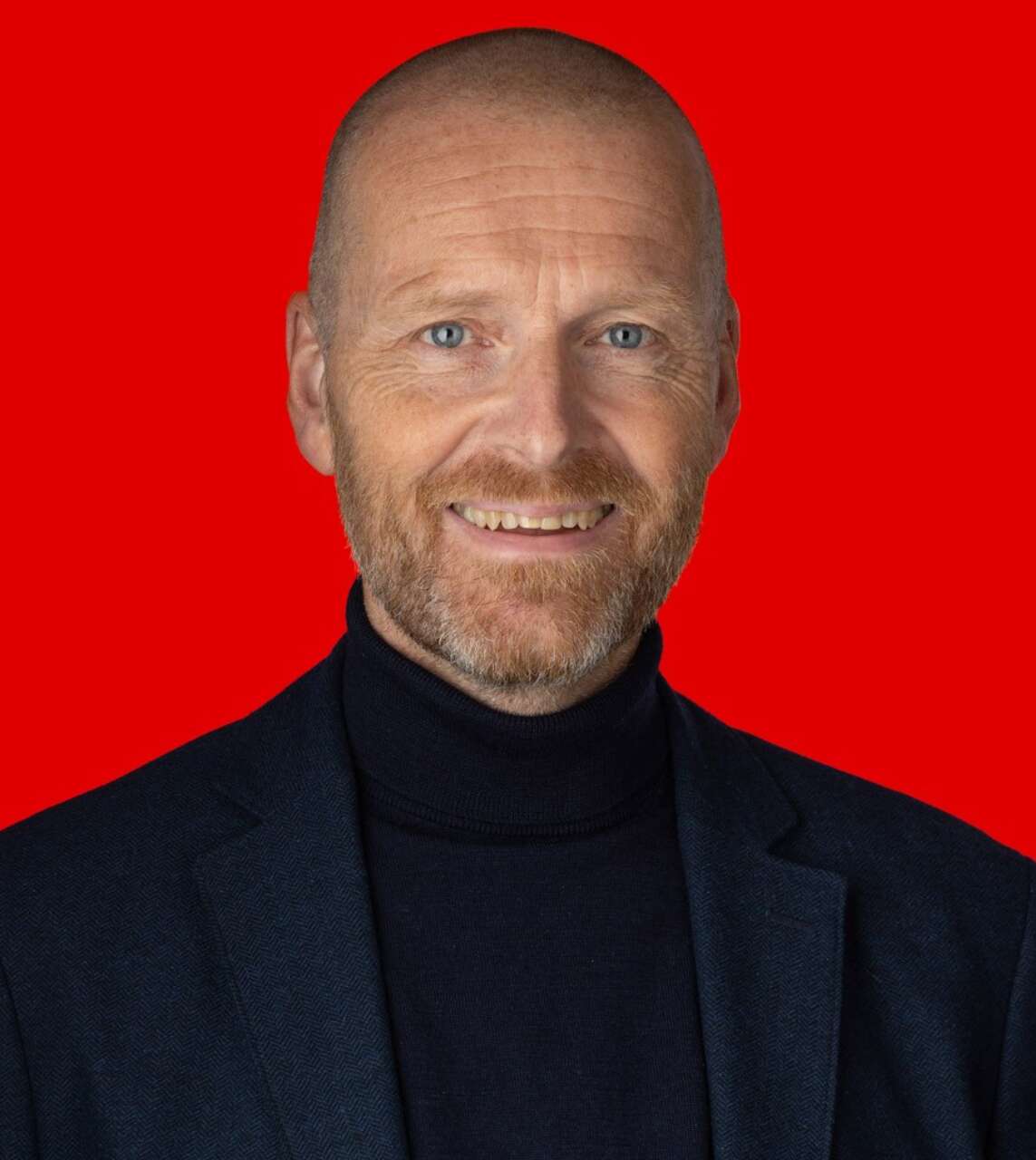 Skaldet mand med skæg, sort rullekrave og jakke, smilende foran rød baggrund.