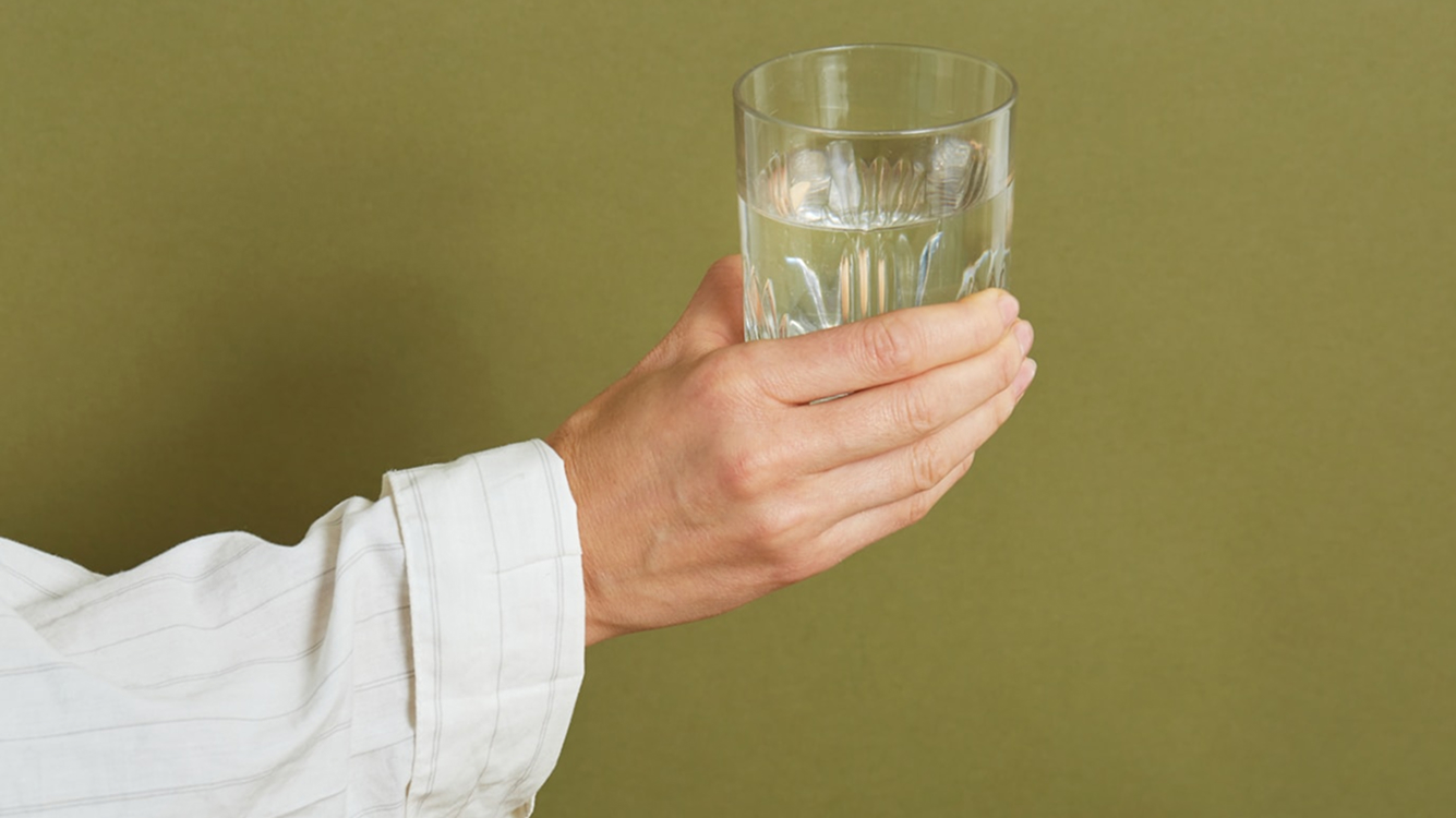 En hand i vit skjorta håller ett glas med vatten mot en grön bakgrund.