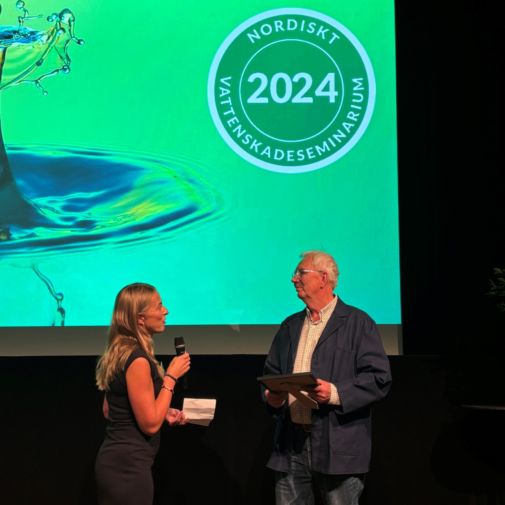 Två personer står på scen framför en grön skärm med texten "Nordiskt Vattenskadeseminarium 2024".
