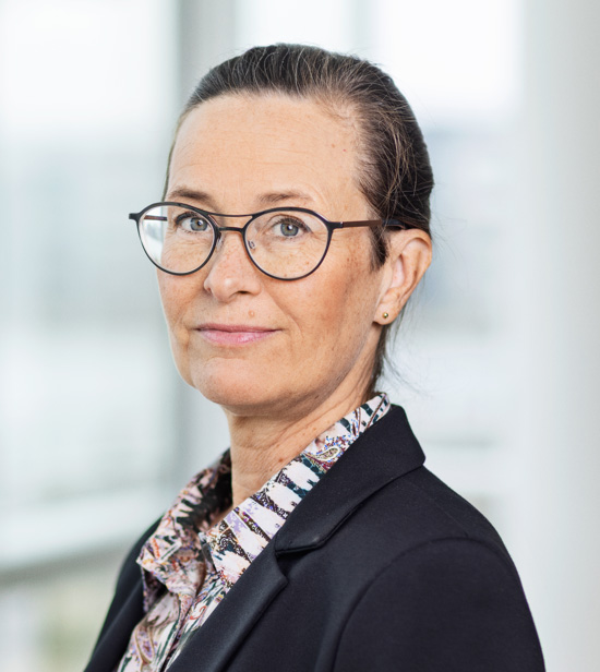 Kvinna med glasögon, uppsatt hår och kavaj tittar in i kameran, ljus bakgrund.