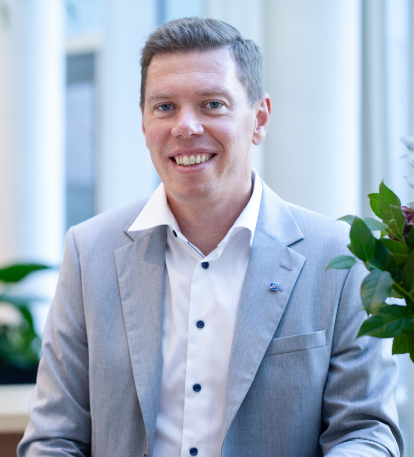 Man i ljusgrå kostym och vit skjorta ler, suddig bakgrund med gröna växter och ljusa pelare.