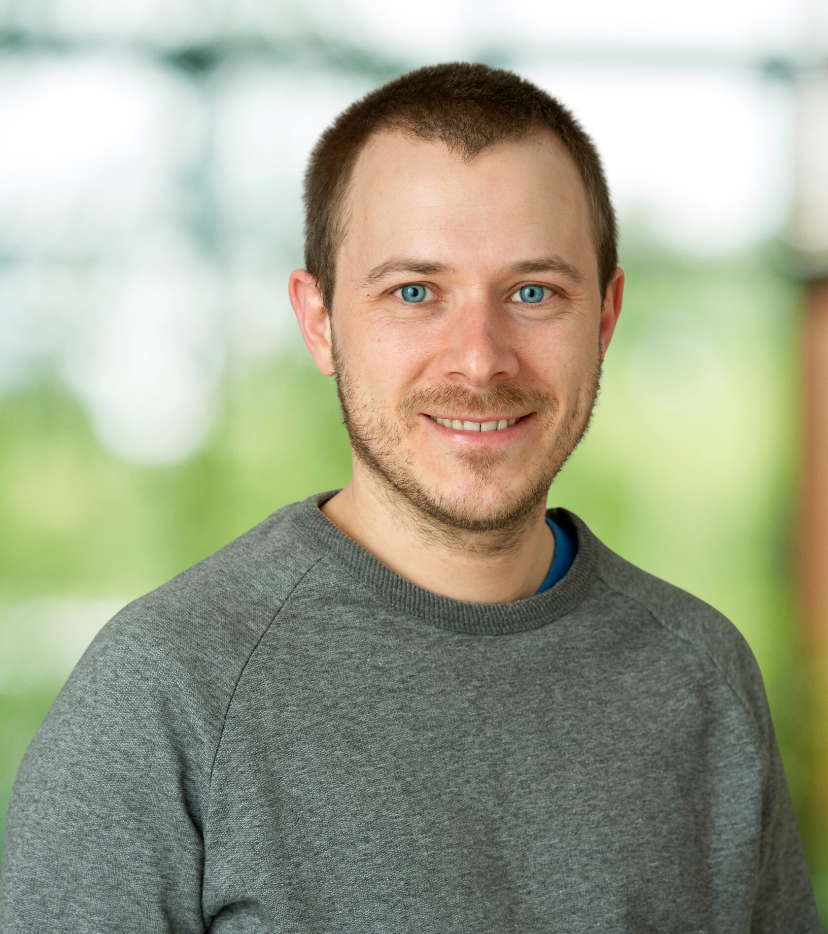 Man med kort brunt hår och blå ögon i grå tröja, ler framför suddig grön bakgrund.