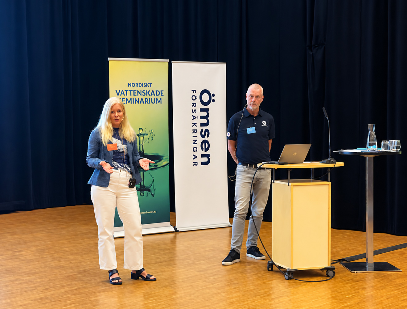 Kvinna och man håller föredrag vid seminarium med rollups, dator och vattenkaraff på scenen.