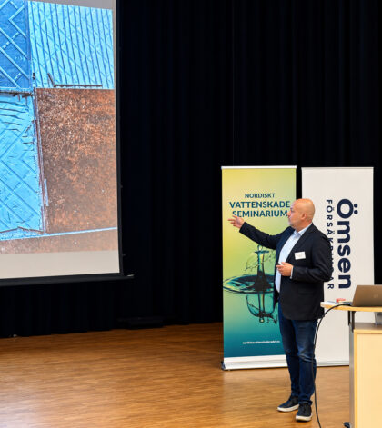 Man ger presentation, pekar mot stor bild på väggen. Banderoller med texten "Vattenskadeseminarium" synliga.
