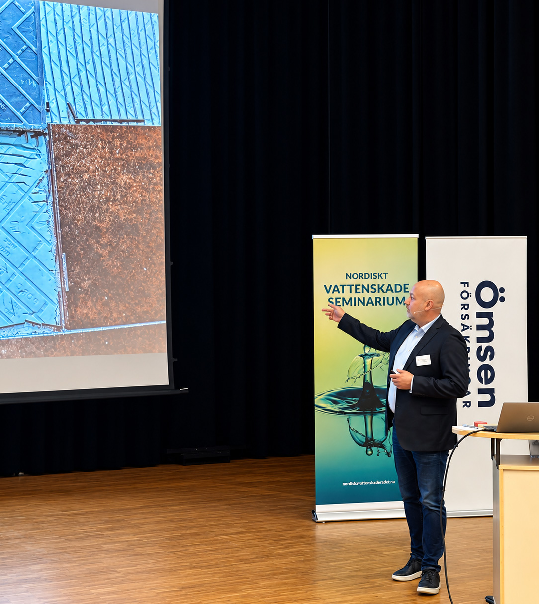 Man ger presentation, pekar mot stor bild på väggen. Banderoller med texten "Vattenskadeseminarium" synliga.