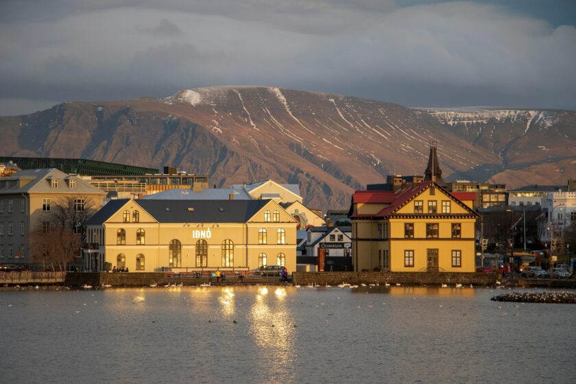 Två hus vid vattnet i Reykjavik, Island, med berg i bakgrunden och solen reflekterande i sjön.